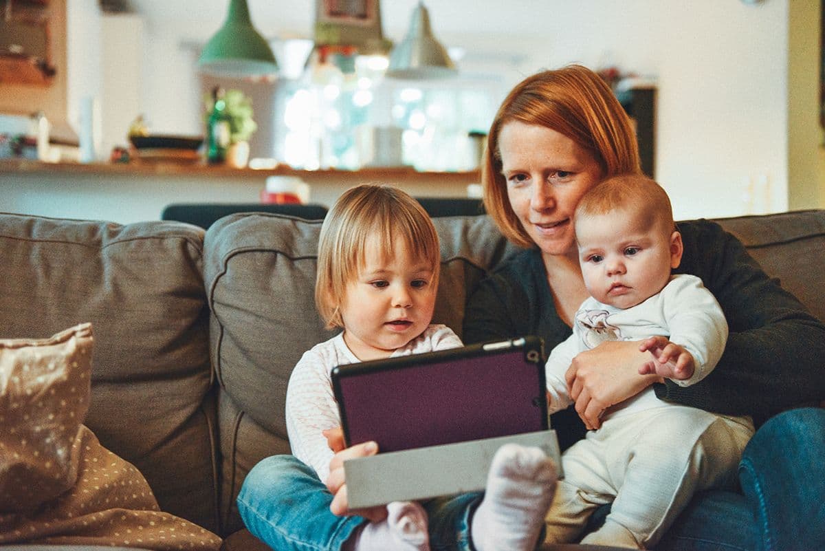 Mom on the couch holding her baby while her toddler watches a tablet